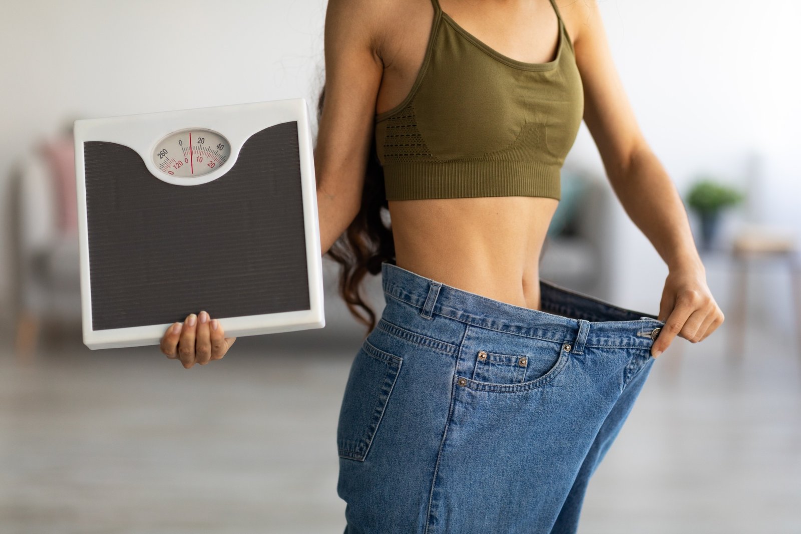 Closeup of Indian woman in oversized jeans holding scales, showing results of weight loss program or Home closeup of indian woman in oversized jeans holding scales showing results of weight loss program or